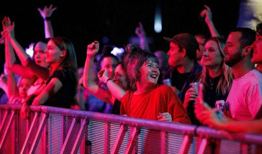 Audience members gather behind a barrier at a live event illuminated by pink and purple stage lighting.