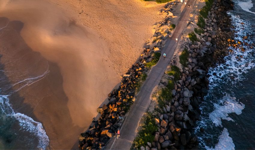 An aerial view of a section of Nobbys Breakwall, with the golden sands of Nobbys Beach to left and waves crashing to the right.