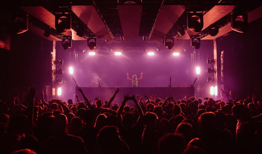DJ onstage in front of a packed bandroom dancefloor, with vibrant lighting and people dancing.