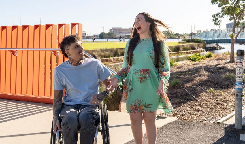 Two visitors, including a wheelchair user, crossing towards a waterfront café along the accessible Honeysuckle promenade with Newcastle Harbour in the background.