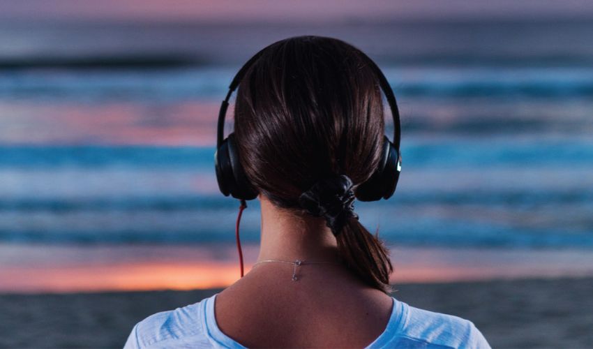 A person wearing headphones looks out toward the ocean at dusk from a beach shoreline.