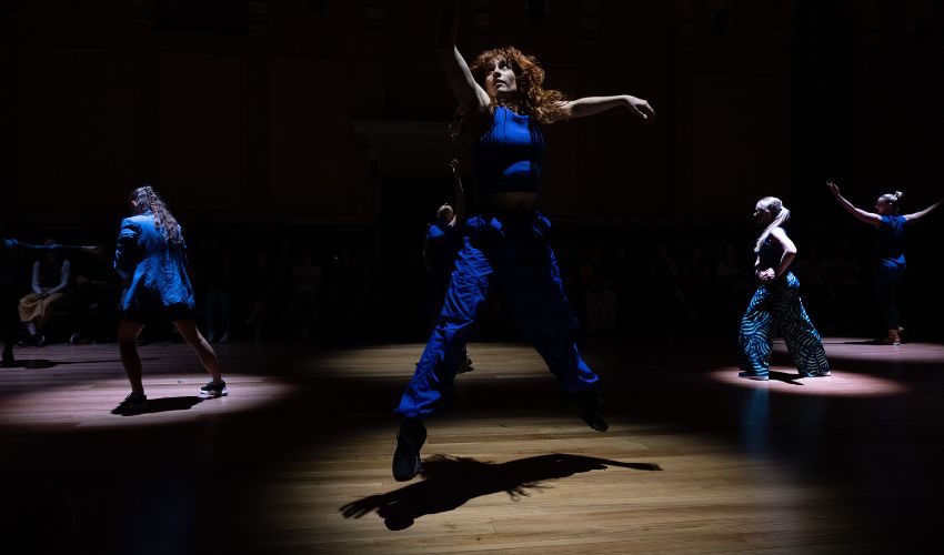 A performer jumps into a spotlight on a dark stage while other performers move in pools of light around the room.