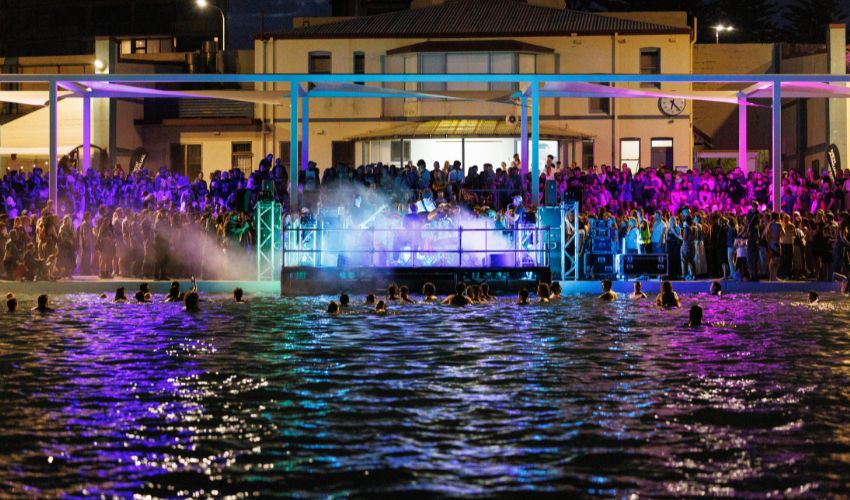 A crowd gathers around a brightly lit poolside stage at night while people swim in the water in the foreground.