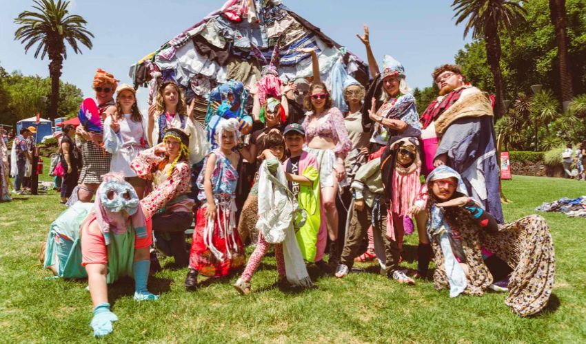 A group of people in colourful handmade costumes pose together on a grassy lawn during an outdoor festival.