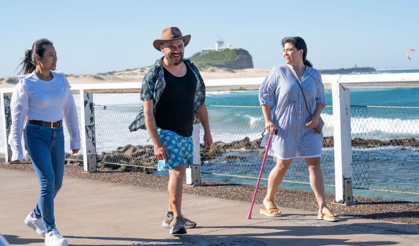 Three friends walking along Newcastle's coastline with Nobbys Headland and the harbour in the background.