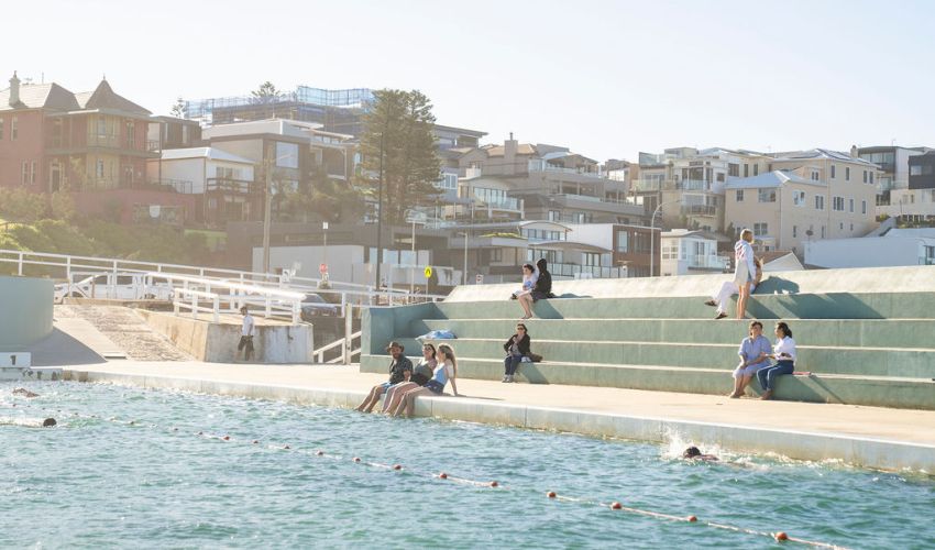 A group of friends sit poolside at the Newcastle Ocean Baths, with the iconic aqua steps behind them.