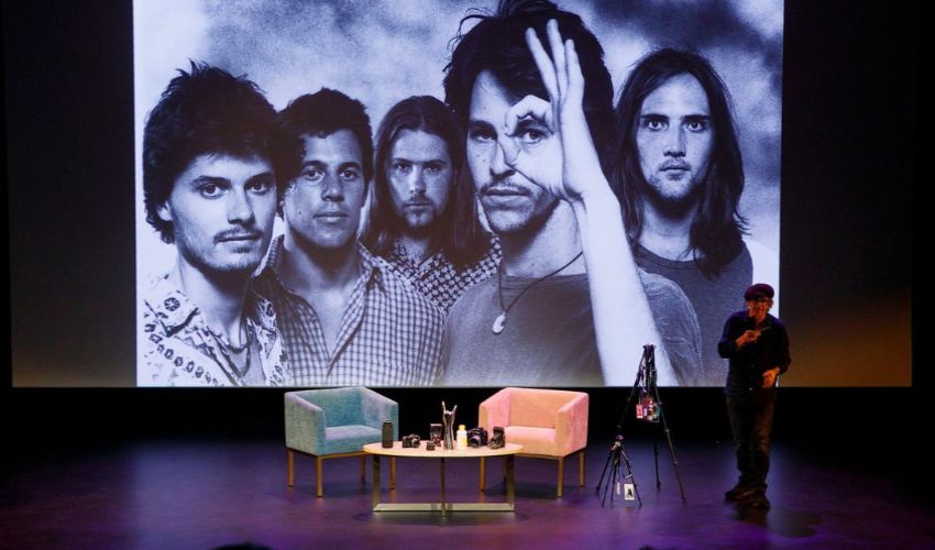 A speaker stands on a theatre stage beside a projector screen displaying a large black-and-white photograph of a band.